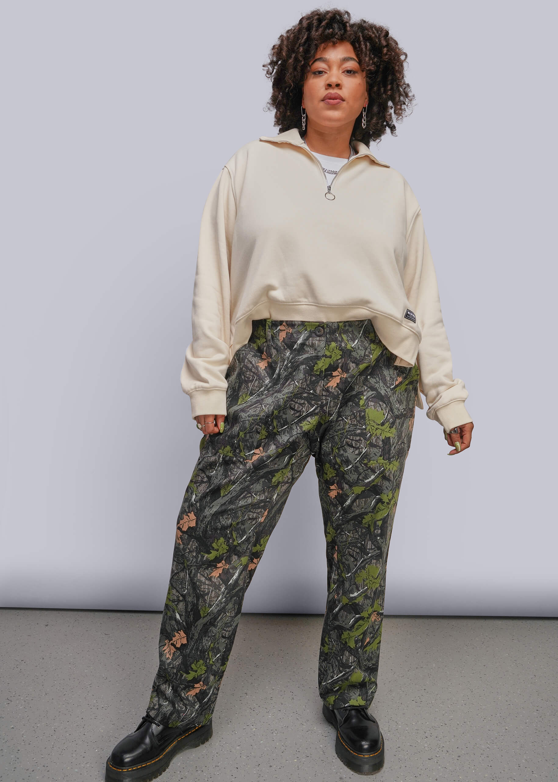 1X=A model stands against a light gray studio background wearing a cream-colored half-zip sweatshirt and dark green camouflage pants with black leather boots.