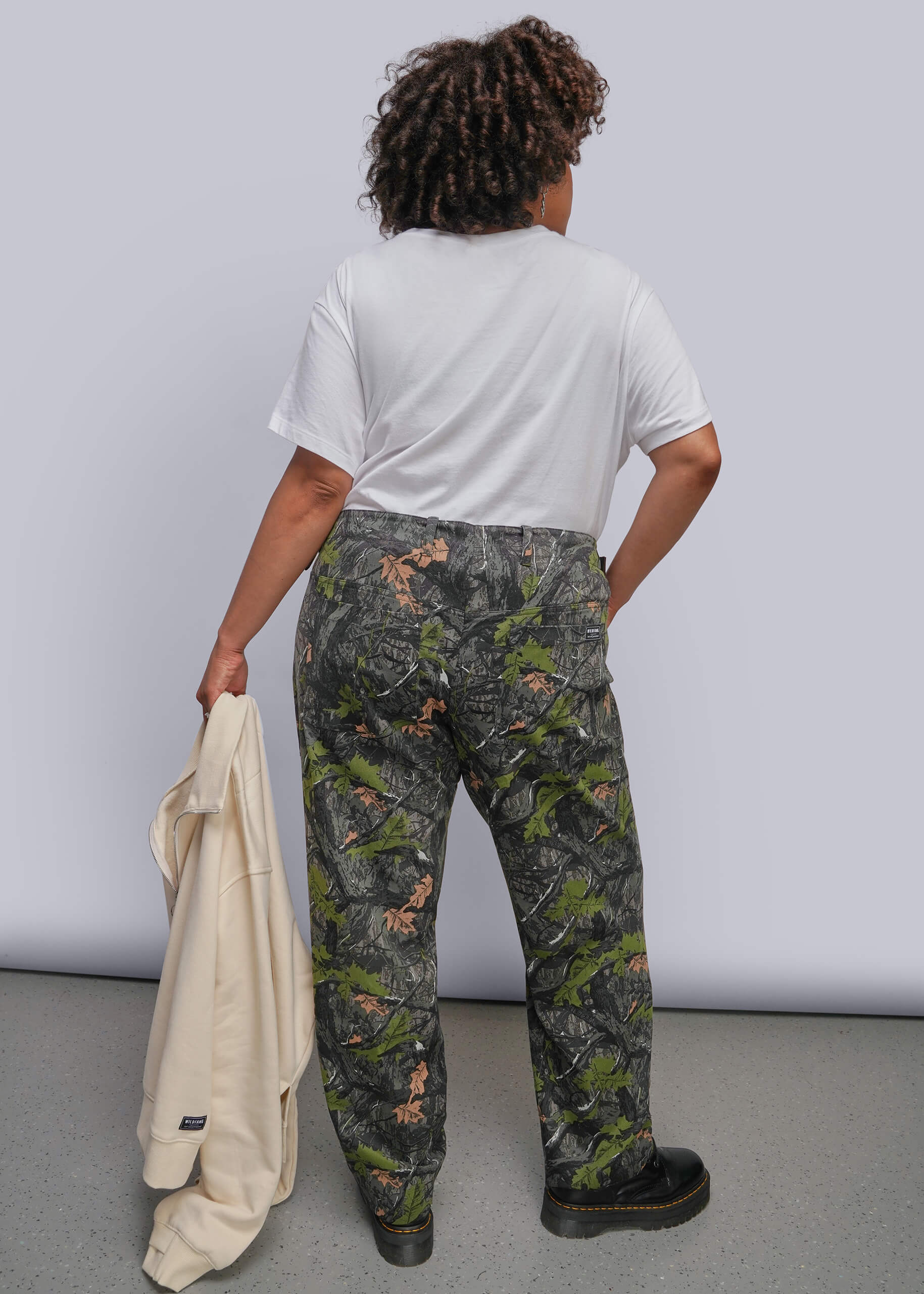 1X=A person shown from the back wearing camouflage pants with a white t-shirt, holding a cream sweatshirt against a light gray studio background.