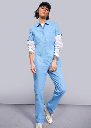 S=Wearing The Essential Coverall in blue stretch cotton over a white shirt and sneakers, a person stands confidently with hands slightly in pockets. Featuring a full-length front zipper, 9 pockets, and a blue cap, the outfit highlights tattoos on a light background.