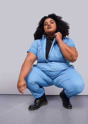 A person with curly hair confidently squats against a white background, wearing The Essential Coverall in light blue, which has a full-length front zipper and is made from stretch cotton. It features 9 pockets for style and functionality and is worn over a black top with black boots.