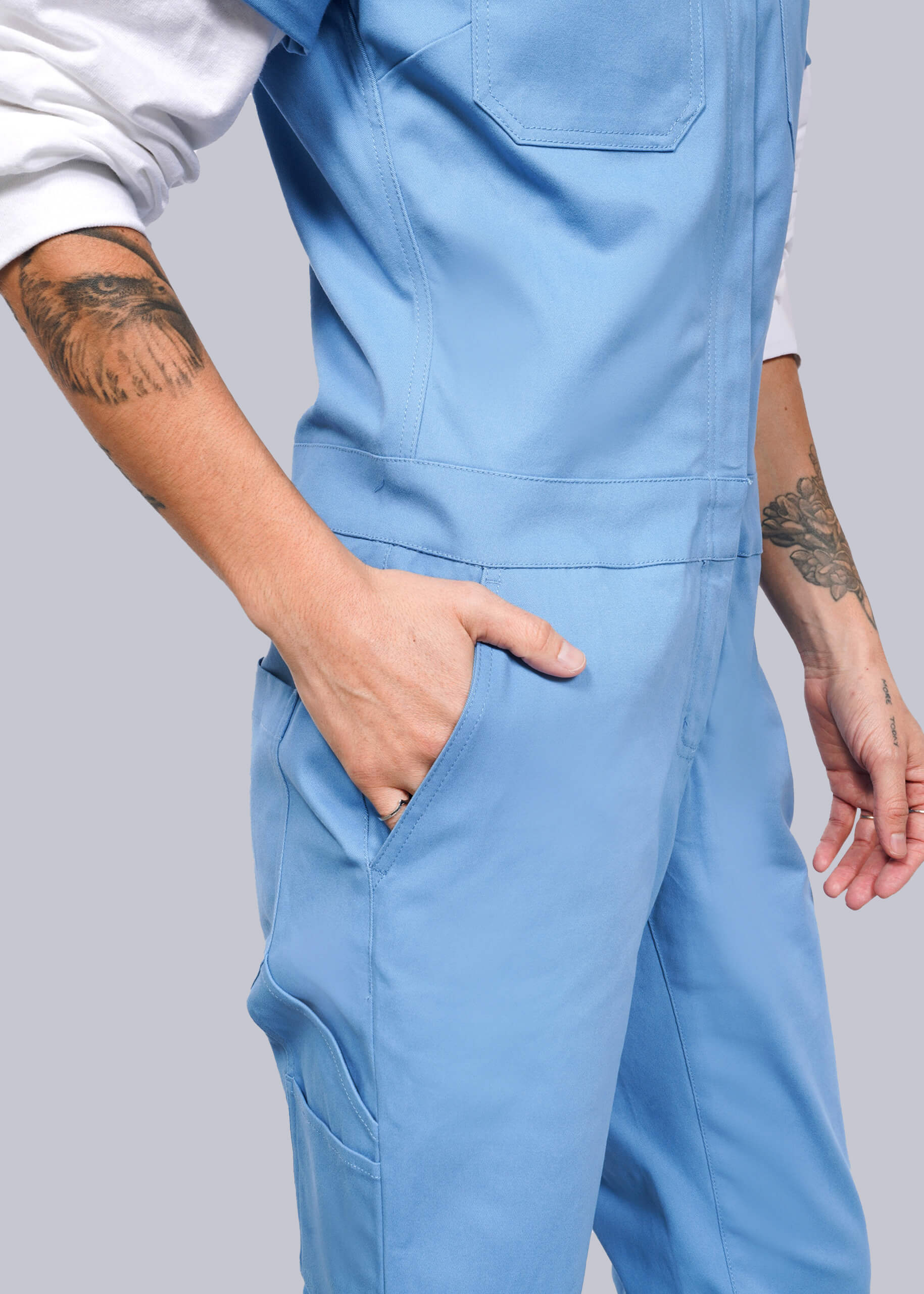 Dressed in The Essential Coverall, a person showcases a blue jumpsuit with a full-length front zipper and stretch cotton fabric. Their tattooed hand rests in their pocket against a plain white background.