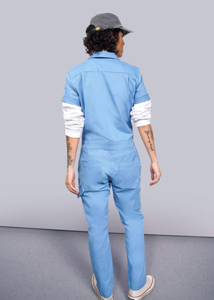 S=A person with curly hair, wearing The Essential Coverall in light blue with a full-length front zipper and a gray cap, faces away from the camera. Tattoos adorn their arms against a neutral backdrop. The stretch cotton of the coverall likely provides comfort and flexibility.