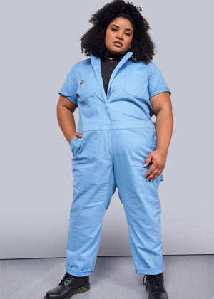 1X=Against a plain backdrop, a person confidently wears The Essential Coverall, a light blue stretch cotton jumpsuit with a full-length front zipper. Paired with black boots and styled curly hair, the outfits 9 pockets offer practicality to their poised look.