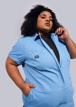 1X=Dressed in The Essential Coverall, a stylish blue stretch cotton jumpsuit with a front zipper and nine convenient pockets, an individual poses confidently against a plain gray backdrop. Their curly hair frames their face as they casually hold the collar with one hand.