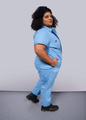 1X=Against a plain white background, a person stands confidently in The Essential Coverall—light blue stretch cotton with a full-length front zipper and nine pockets. With black boots and curly hair adding flair, their hands are casually tucked in.