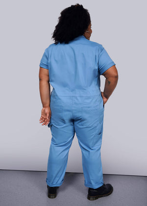 1X=Facing away from the camera with short curly hair, a person wears The Essential Coverall in light blue stretch cotton featuring a full-length front zipper. Theyre paired with black shoes and hold an object in their right hand against a plain white backdrop.
