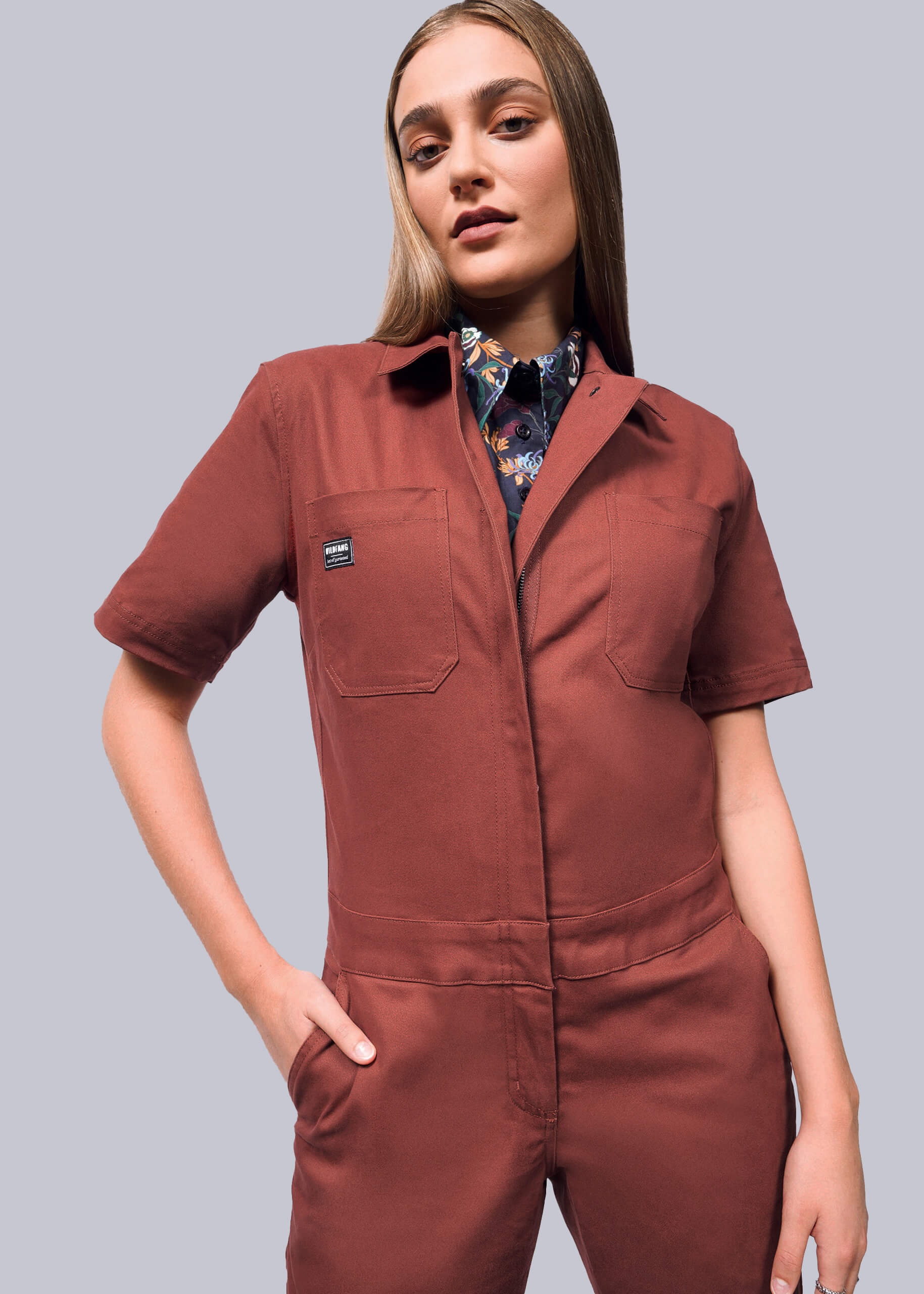 S=A person confidently models The Essential Coverall, a relaxed fit rust-colored jumpsuit featuring short sleeves, a zipper front, and pockets. Its crafted from stretch cotton and layered over a patterned shirt. The background is plain light gray.