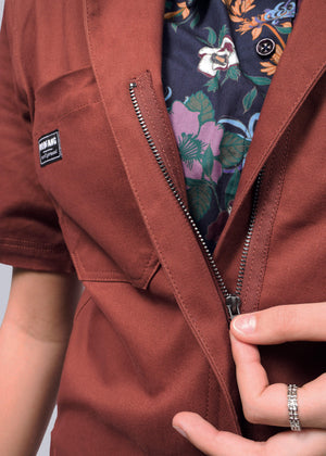 S=Dressed in The Essential Coverall with a rust hue and partially open zipper, revealing a floral patterned shirt underneath, the person enjoys the relaxed fit while holding the zipper. One hand is adorned with rings.