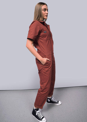 S=Showcasing the relaxed fit of stretch cotton, a person wearing The Essential Coverall—a red-orange short-sleeve jumpsuit—and black sneakers stands with hands in pockets, facing slightly left against a plain light gray background.