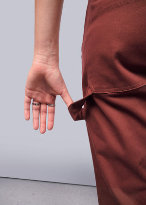 S=A person is shown in rust-colored, relaxed-fit pants, pulling a fabric loop at the waist. Made from stretchy cotton, these pants blend comfort with style. The hand wears two silver rings against a plain background. These are named The Essential Coverall.