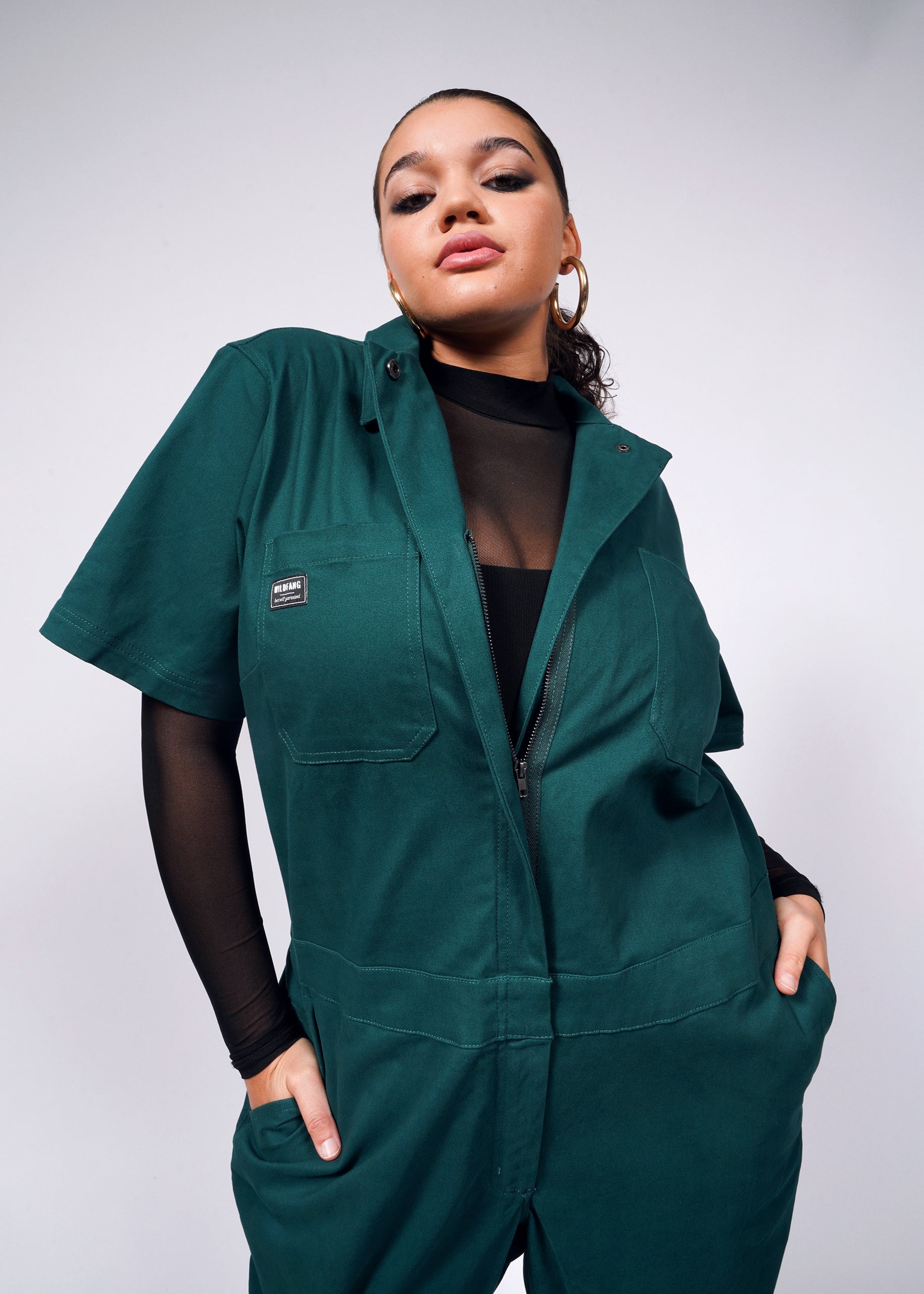 1X=A person poses confidently in The Essential Coverall, made of soft brushed stretch cotton in green, featuring a full-length front zipper and 9 pockets. They wear a black mesh top underneath, hoop earrings, and have slicked-back hair against a plain neutral background.