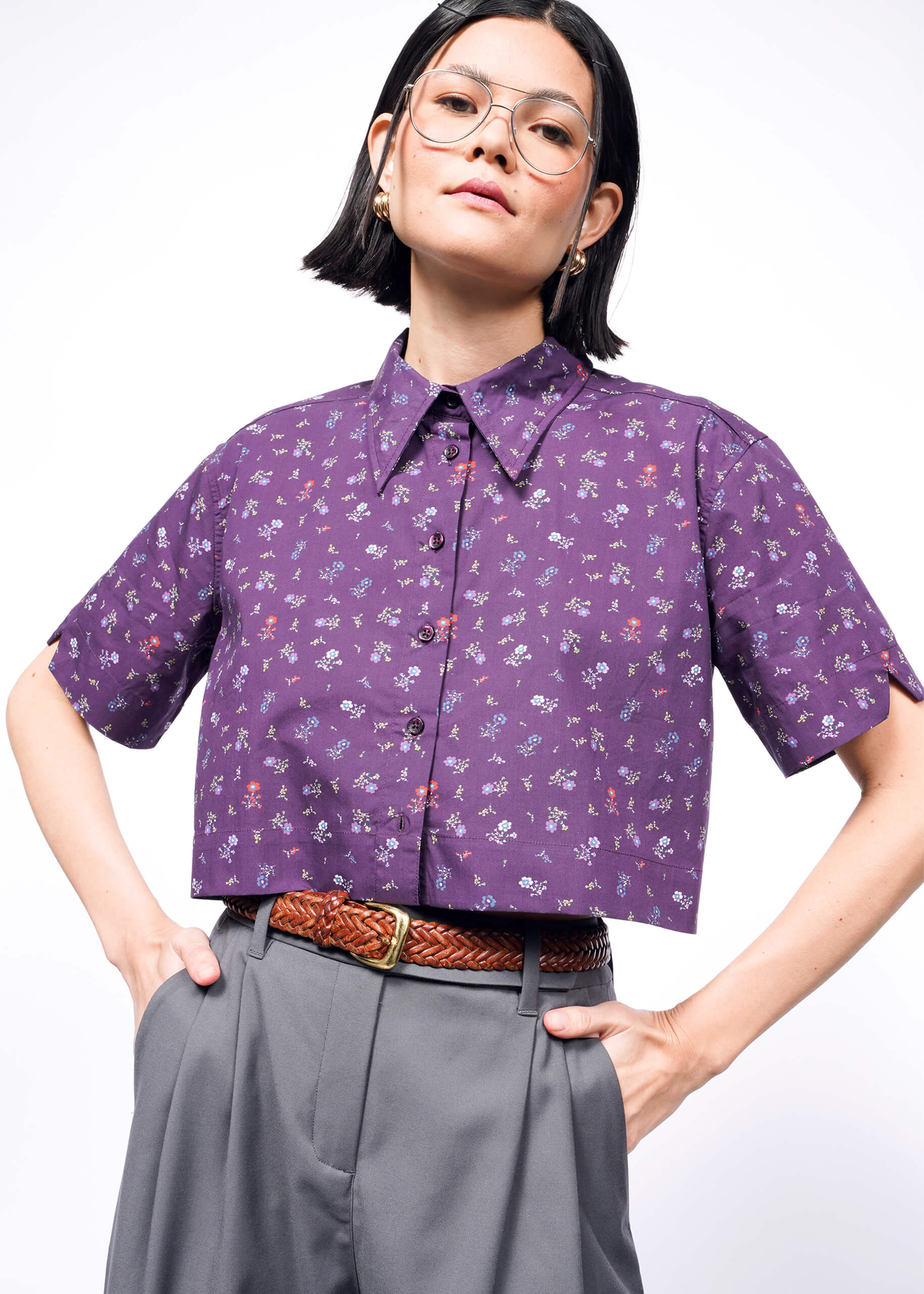 A person wearing glasses and The Essential Cropped Button Up in a purple floral cotton print, with Wildfang buttons, paired with high-waisted gray trousers. The oversized fit exudes confidence as they pose against a white background, hands in pockets.