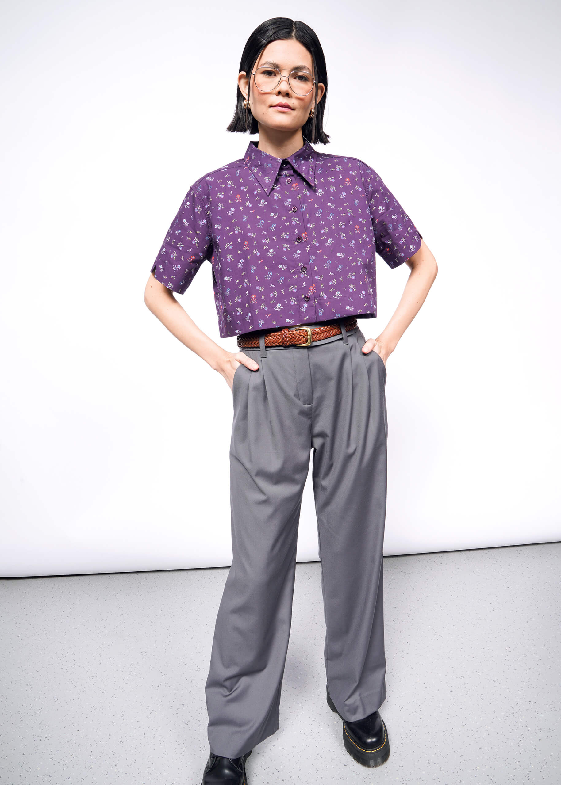 A person with short black hair and glasses confidently stands against a plain wall, wearing an oversized purple Essential Cropped Button Up featuring Wildfang branded buttons. They pair it stylishly with gray high-waisted pants, a brown belt, and black shoes, hands placed on hips.