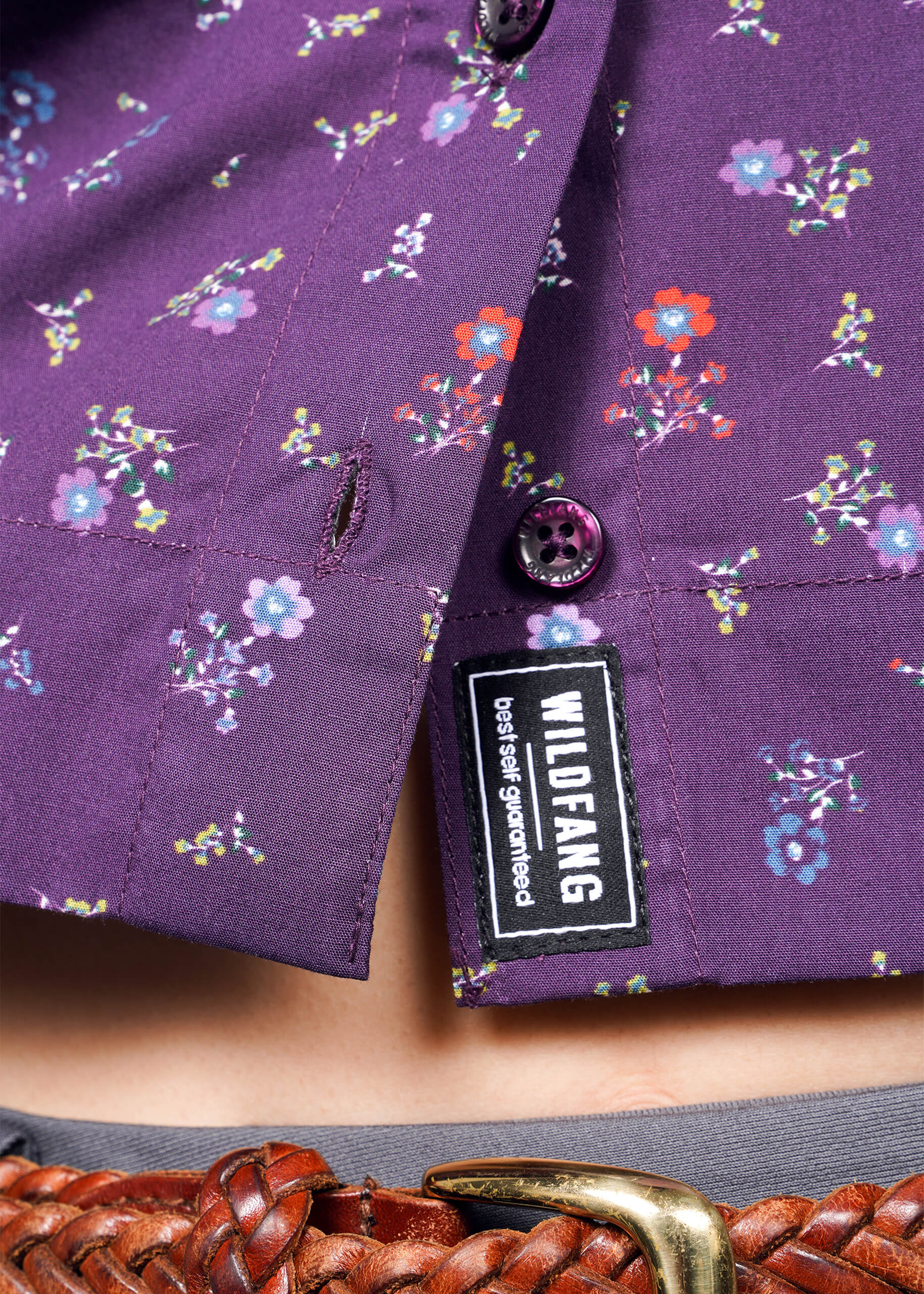 Close-up of The Essential Cropped Button Up, a purple cotton shirt with small floral patterns and Wildfang branded buttons. The oversized fit is tucked into pants with a brown woven belt, featuring a visible Wildfang tag.