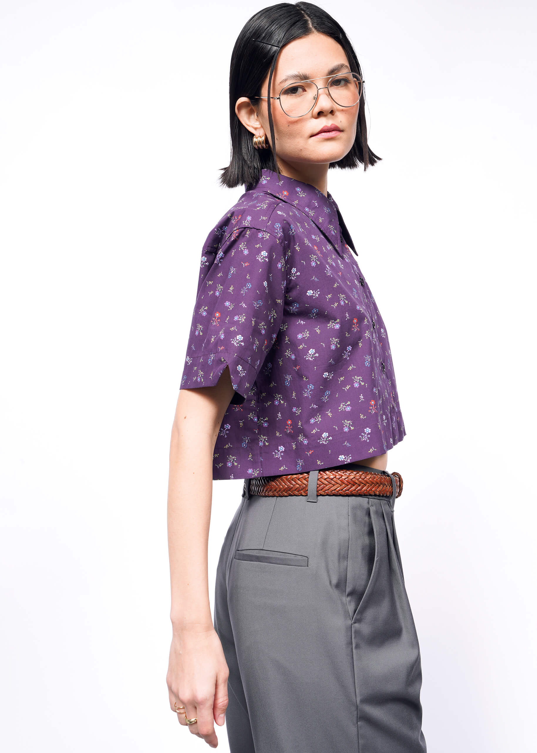 A person with shoulder-length black hair wearing glasses stands in profile, dressed in The Essential Cropped Button Up by Wildfang, a purple floral cotton shirt paired with gray pants and a brown braided belt. Their neutral expression stands out against the white background.