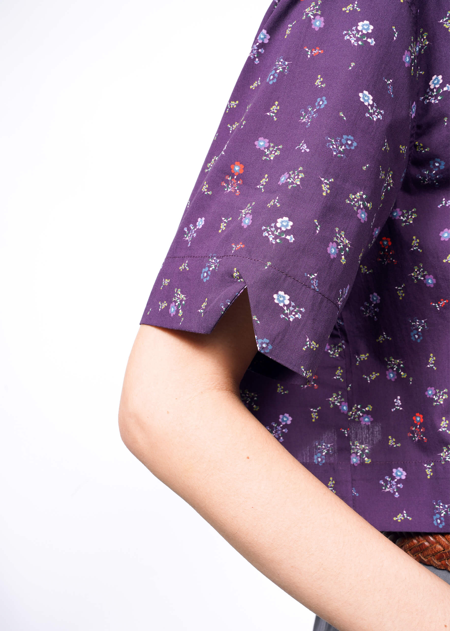 A person wears The Essential Cropped Button Up, a purple short-sleeved top adorned with small, colorful floral patterns. The fabric has a sleeve notch, and the oversized fit ensures comfort. Wildfang-branded buttons accentuate the style against the white backdrop.