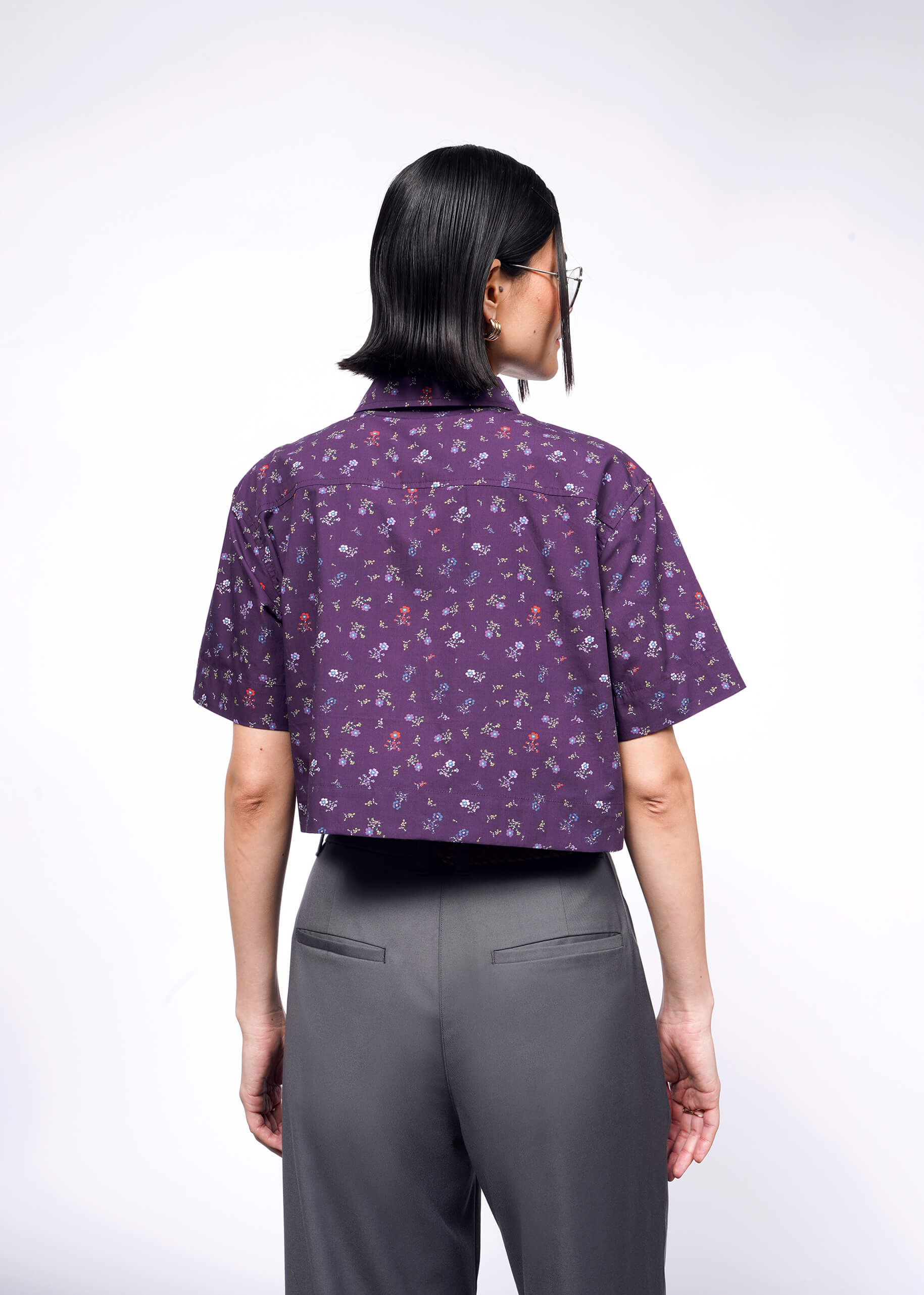 A person with dark hair stands with their back to the camera, wearing The Essential Cropped Button Up, a purple cotton shirt with short sleeves, a small floral pattern, and Wildfang branded buttons. They pair it with gray pants against a plain white backdrop.