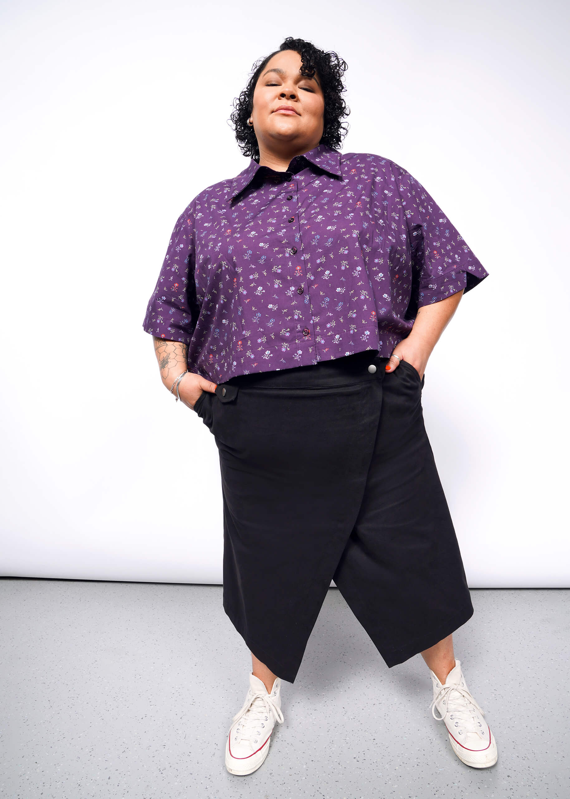 A person stands confidently with hands in pockets, wearing the purple floral-patterned The Essential Cropped Button Up featuring Wildfang-branded buttons. Paired with black wide-leg pants and white sneakers, the oversized fit is captured against a plain background from a slightly low-angle perspective.