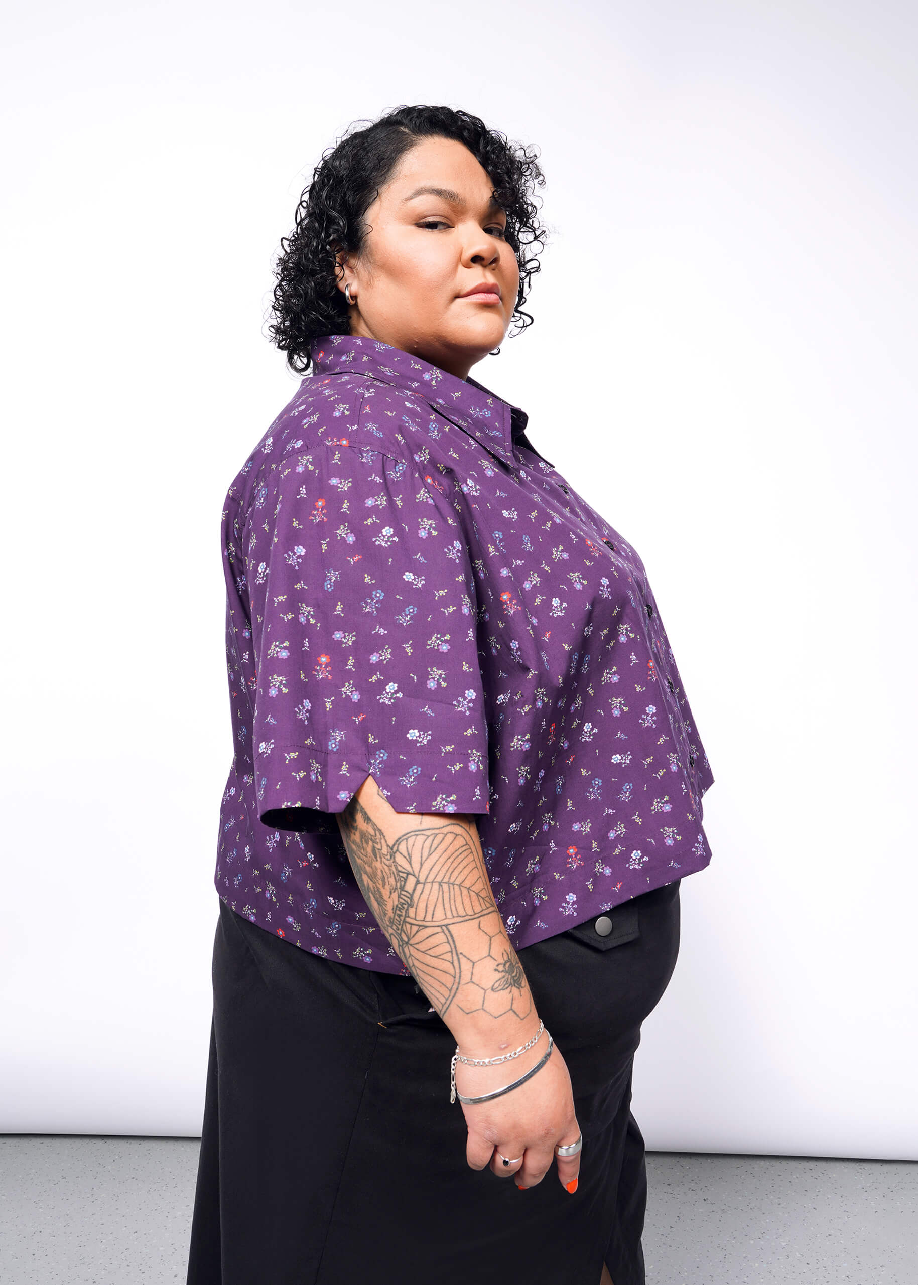 A person with curly hair confidently poses in The Essential Cropped Button Up, a purple floral cotton shirt featuring Wildfang branded buttons, paired with a black skirt. They showcase a forearm tattoo and bracelets against a plain white wall.