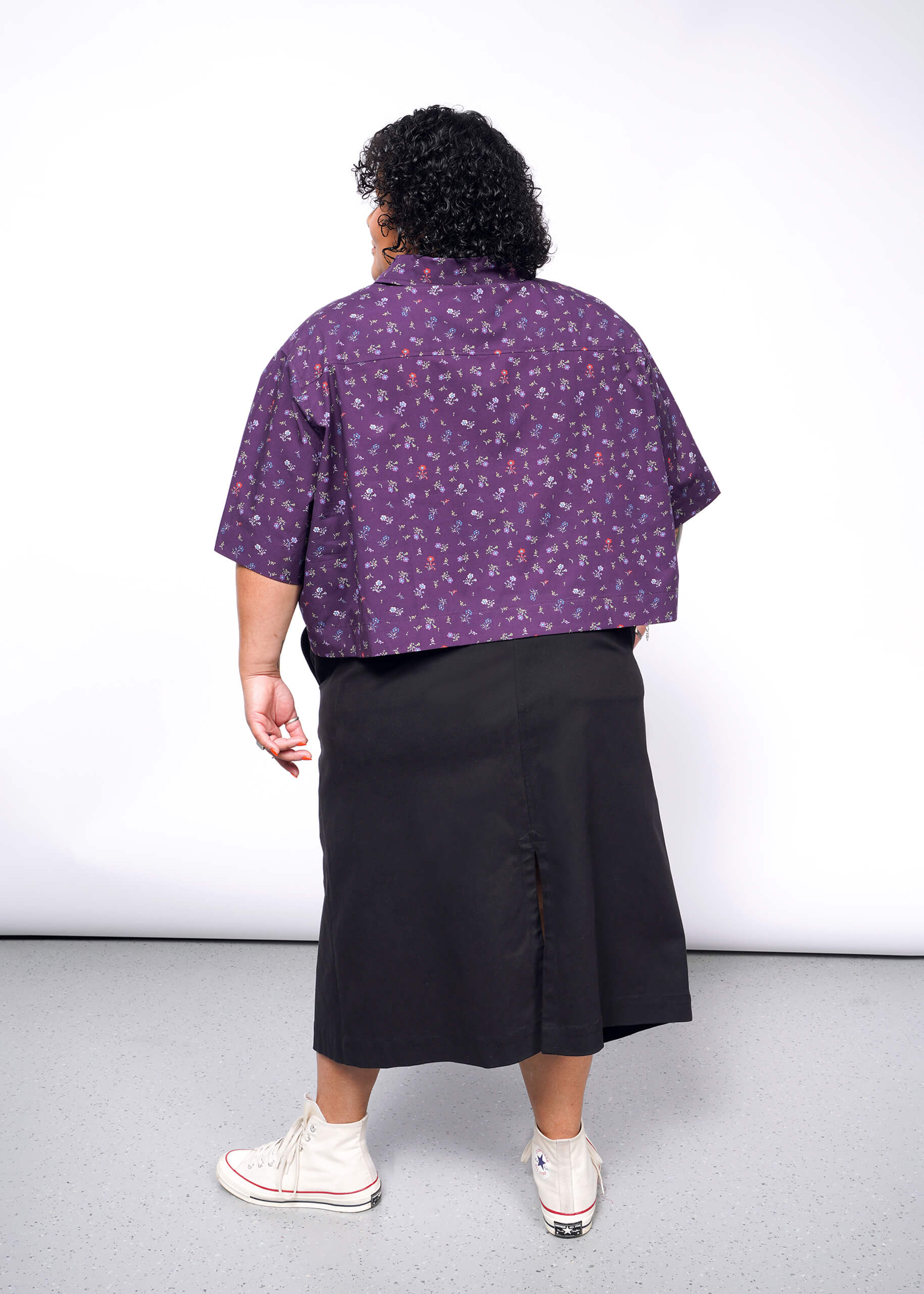 Against a plain white wall, a person with curly hair wears The Essential Cropped Button Up in purple with patterns and a black skirt. The look is completed by white high-top sneakers, with Wildfang branded buttons adding a unique touch.