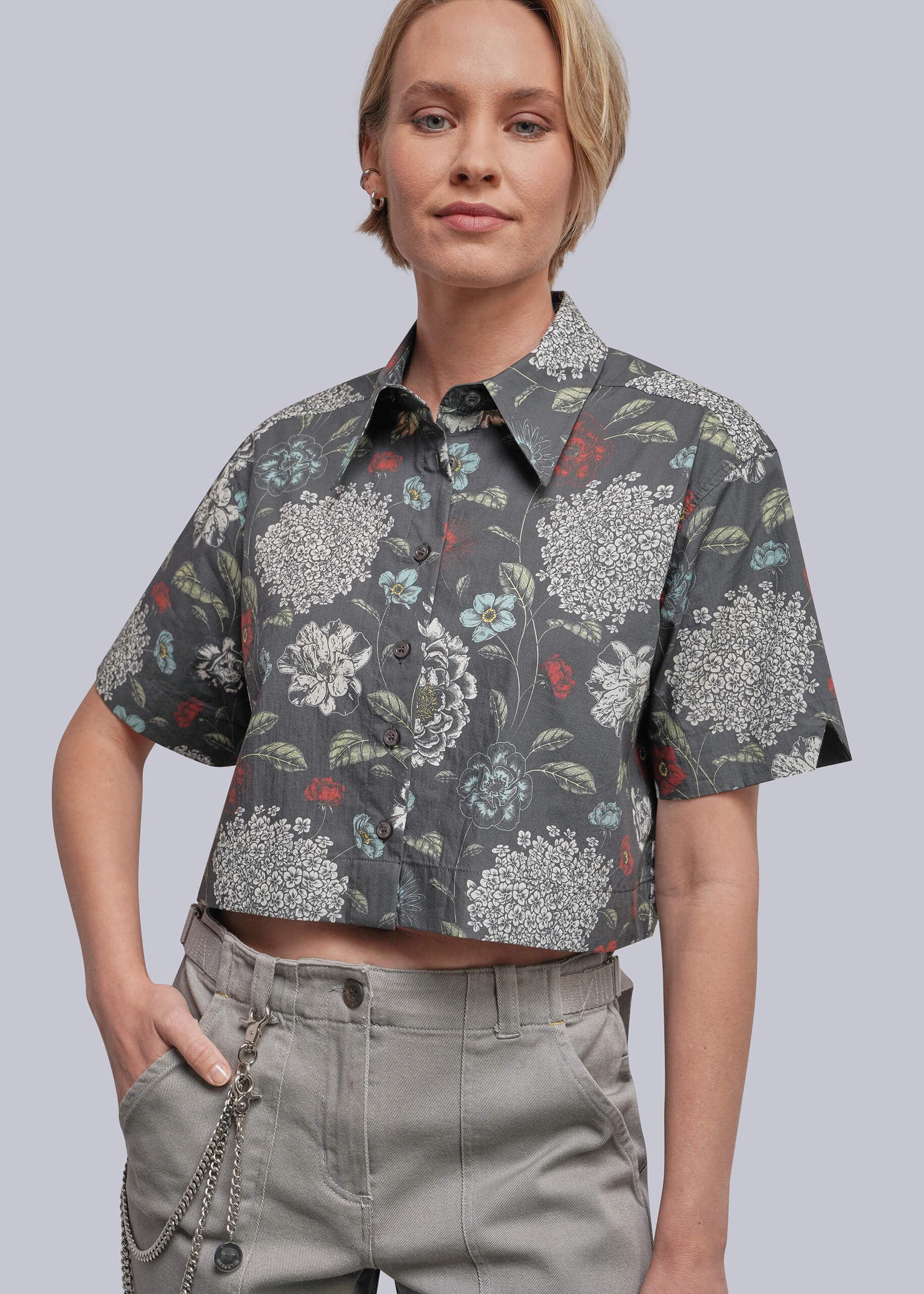 A woman wearing a cropped short-sleeve button-up shirt with a dark gray floral print and light gray pants, photographed against a plain light gray studio background.