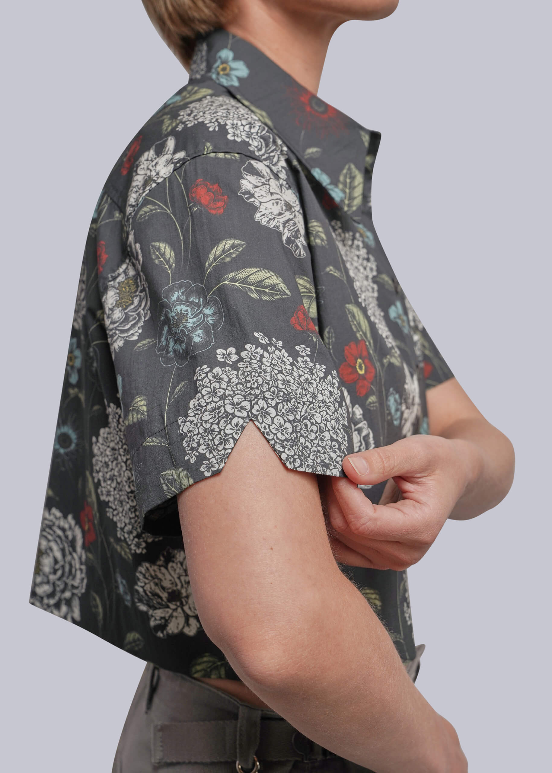 A close-up side view of a person wearing a short-sleeve button-up shirt with a dark gray base and a dense white, red, and green floral print. The person is adjusting the cuff of the shirt against a solid light gray background.