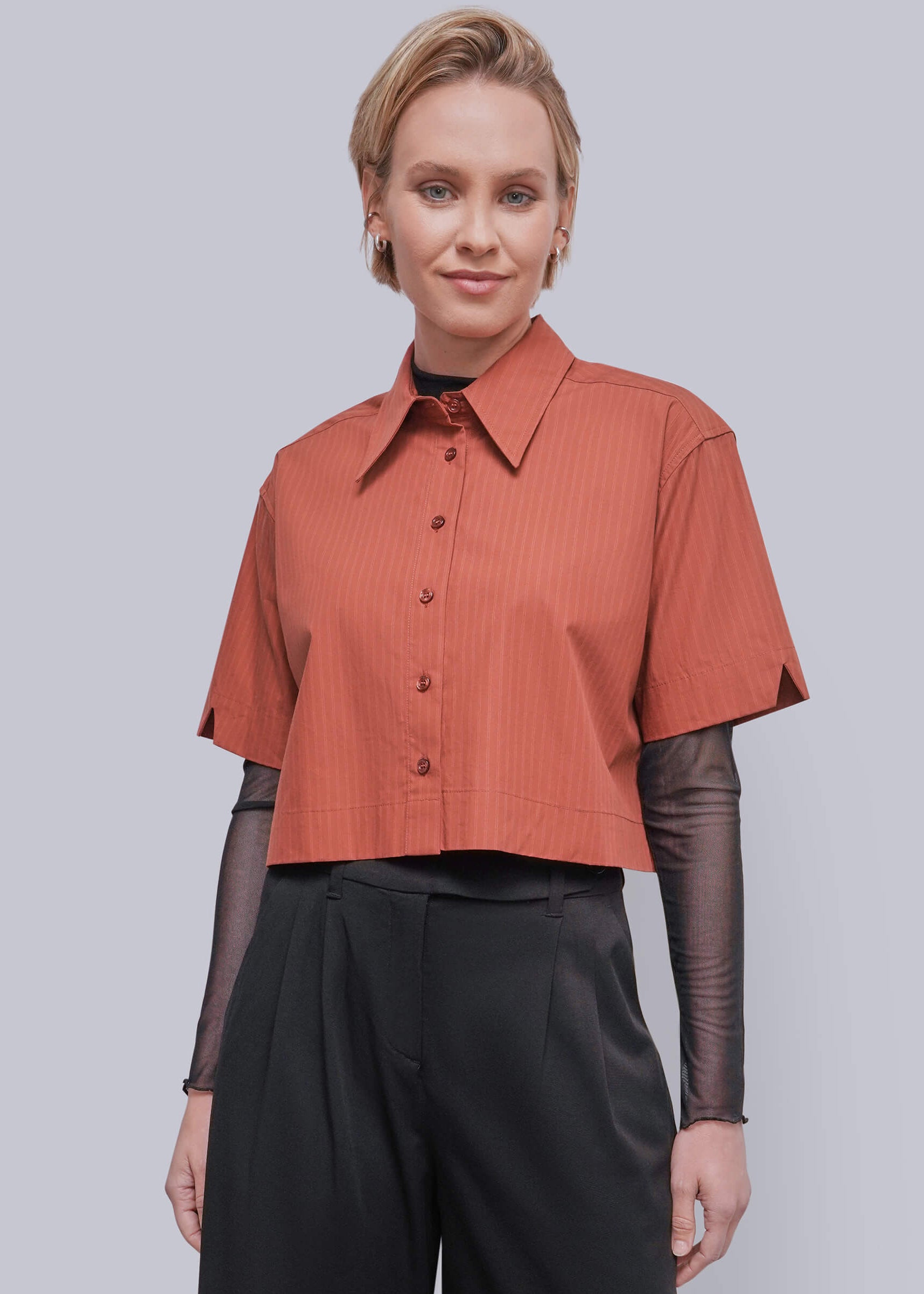 S=A woman modeling a short-sleeve cropped button-up shirt in a muted terracotta or burnt orange color with subtle vertical texture, layered over sheer black long-sleeve sleeves and black trousers, against a solid light gray studio background.
