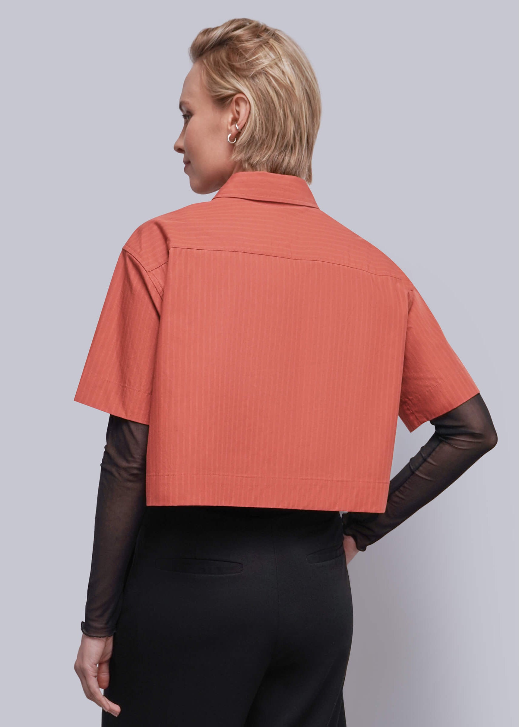 S=A woman shown from the back wearing a short-sleeve cropped collared shirt in a muted coral-orange color with subtle vertical texture, layered over sheer black long sleeves and black trousers, against a solid light gray studio background.