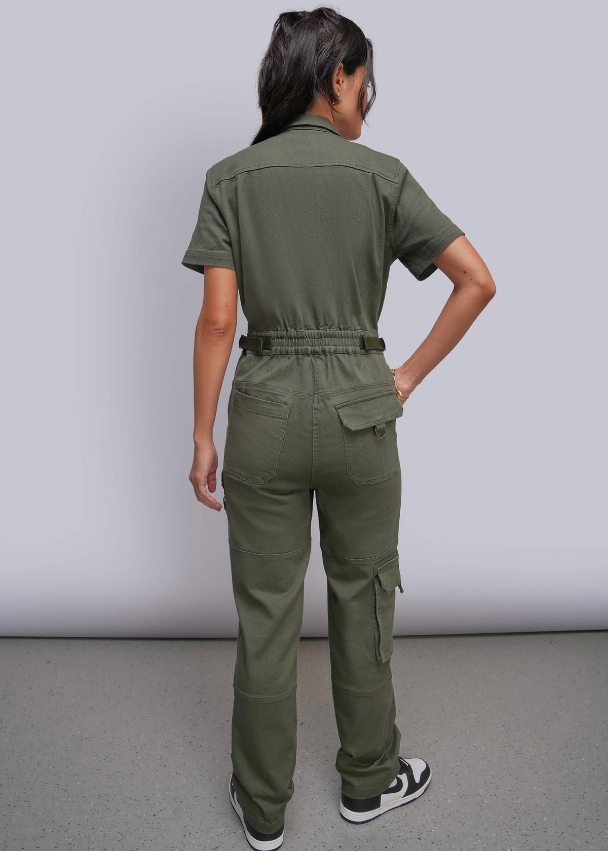 S=A woman shown from the back wearing an olive green short-sleeve utility jumpsuit with cargo pockets and an adjustable waist, paired with black and white sneakers, against a light gray studio background.