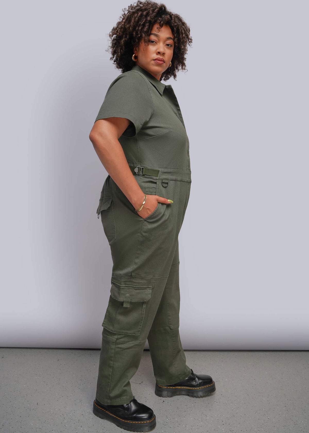 1X=A woman modeling an olive green short-sleeve utility jumpsuit with cargo pockets and a belt, paired with black platform shoes, against a light gray studio background.