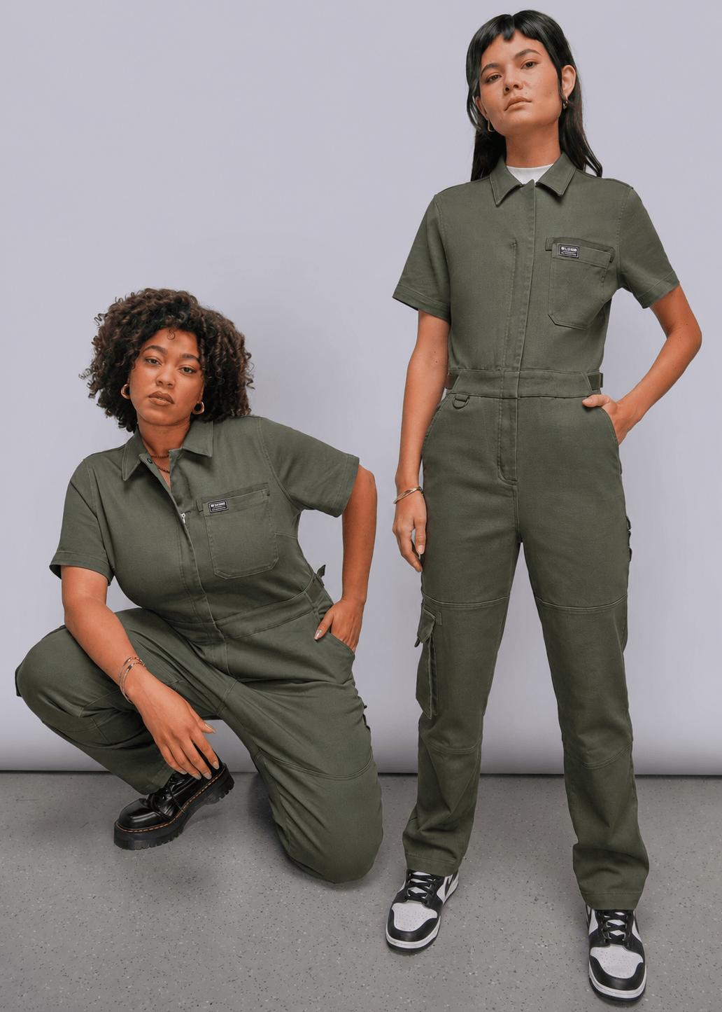 Two models wearing matching olive green short-sleeve utility jumpsuits, posed against a neutral light gray studio background.