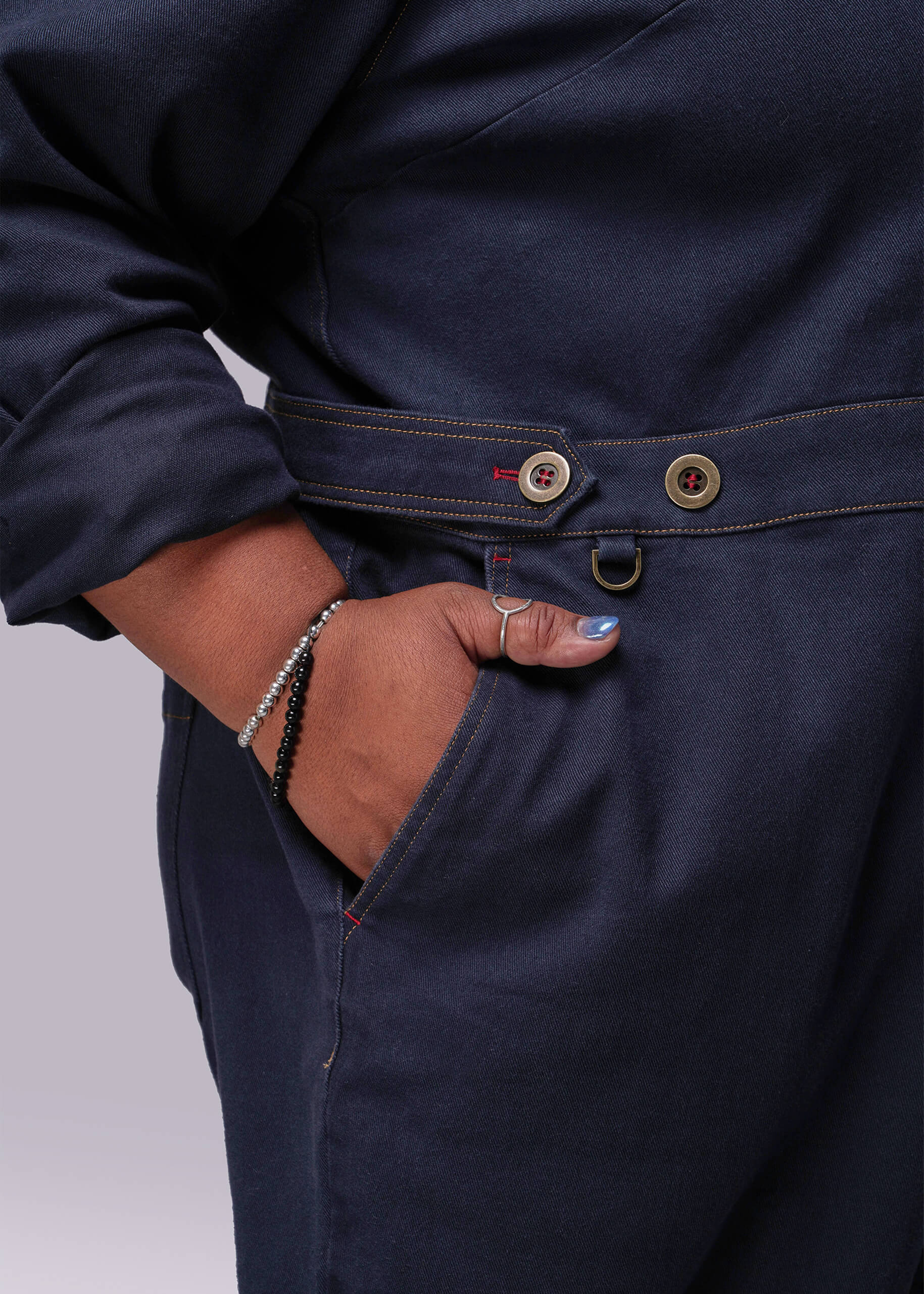 1X=Navy blue jumpsuit with buttons and a person's hand in the pocket on a light gray background