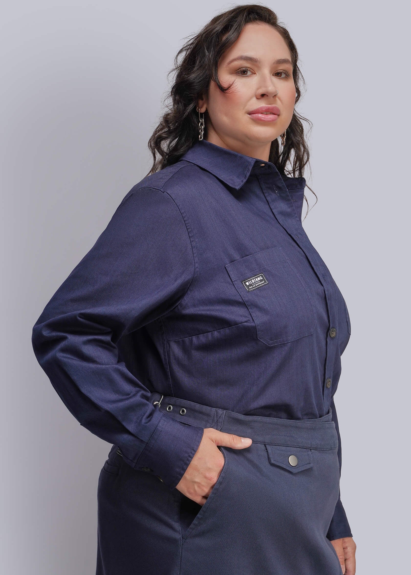 1X=A woman wearing a dark navy blue long-sleeve button-up shirt with a chest pocket and a matching dark skirt, posed against a solid light gray studio background.