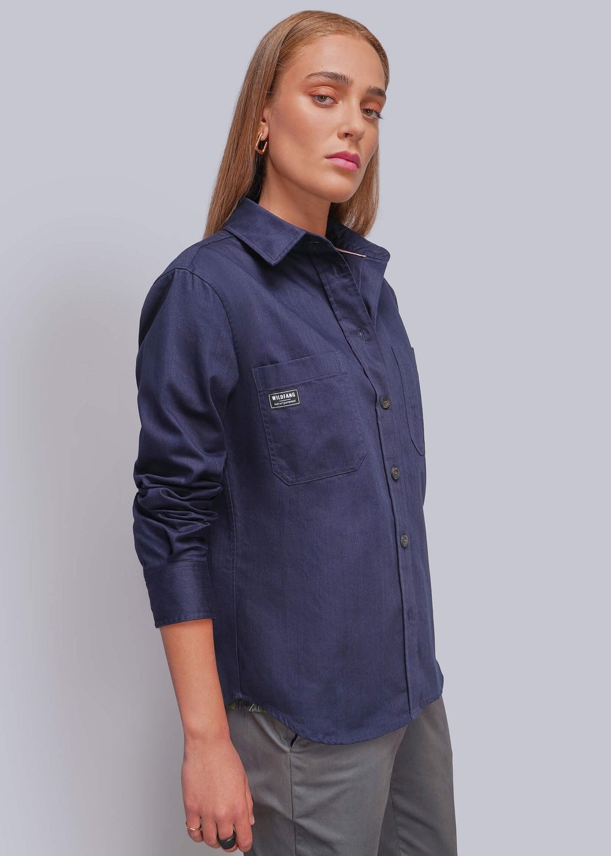 S=A woman modeling a navy blue long-sleeve button-up shirt with a chest pocket, paired with gray trousers, against a solid light gray background.