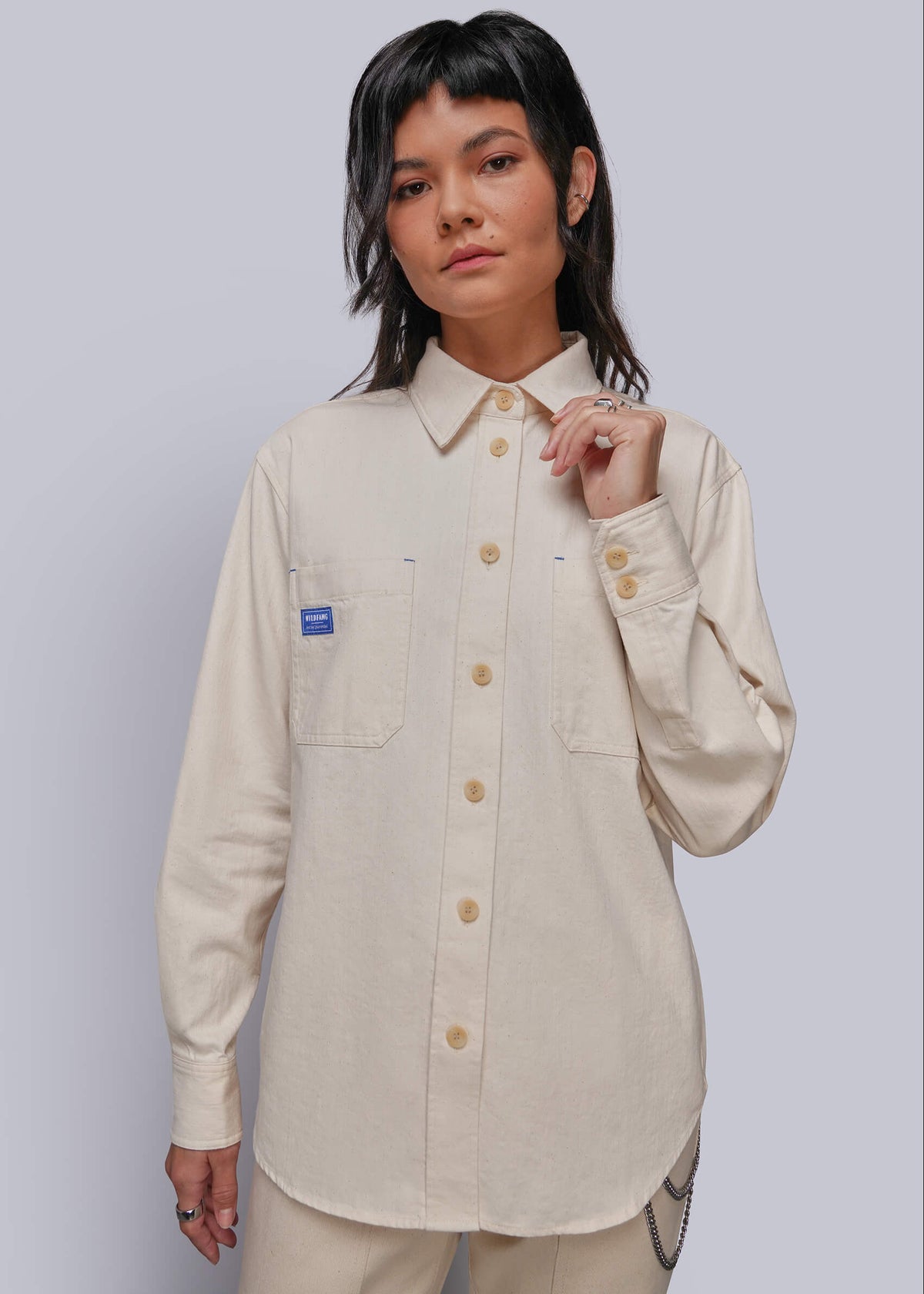 S=A model wearing a light beige long-sleeve button-up shirt with a pointed collar, chest pockets, and a small blue brand patch on the left pocket, paired with matching beige pants, against a solid light gray background.