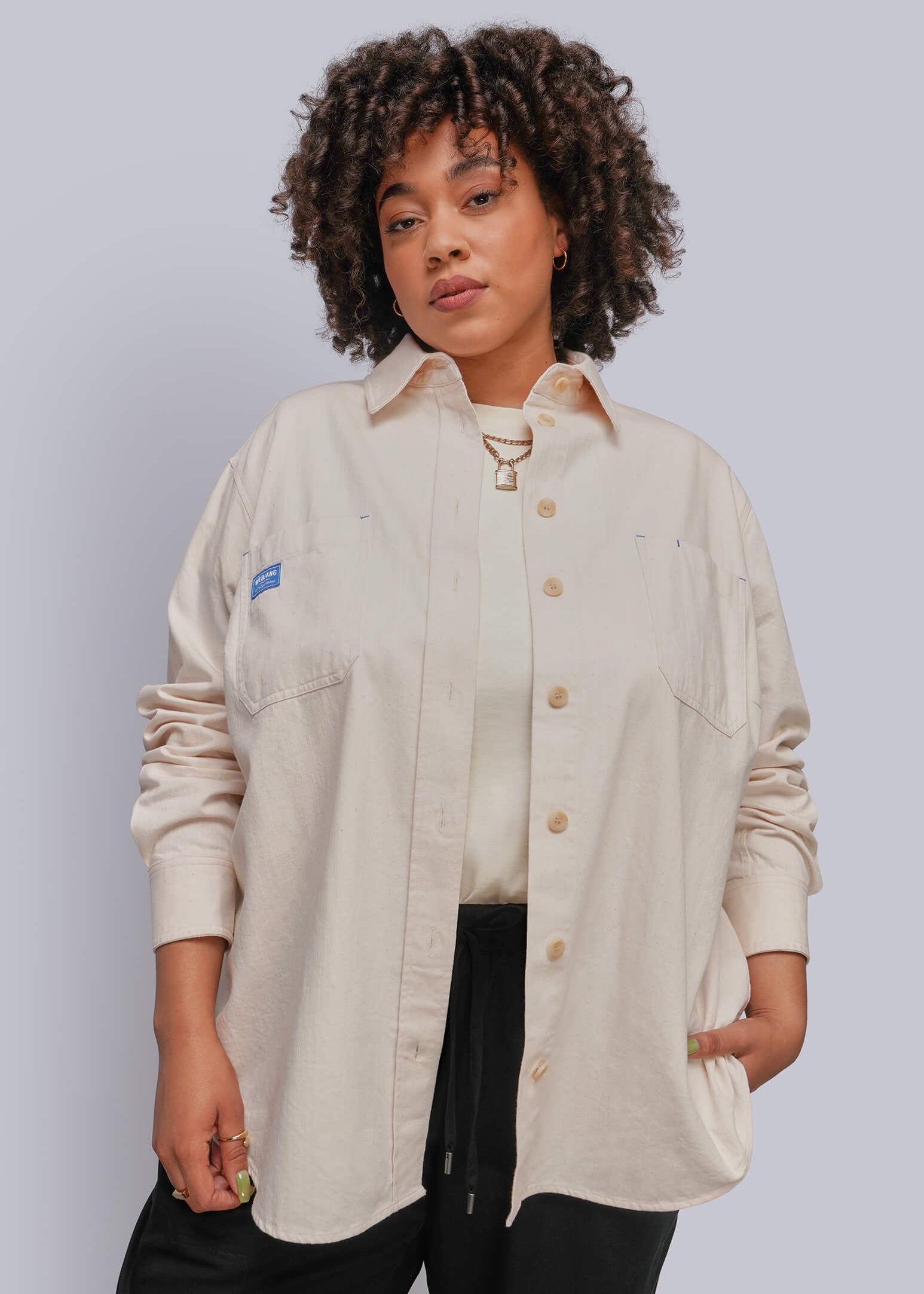 1X=A woman with curly hair wearing a light beige button-up shirt with a small blue logo patch on the chest pocket, paired with black pants, against a solid light gray background.