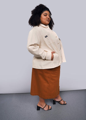 1X=A person with curly hair confidently wears The Essential Denim Utility Jacket with deep pockets, paired with a brown skirt and black heeled sandals, standing in a studio against a neutral background.