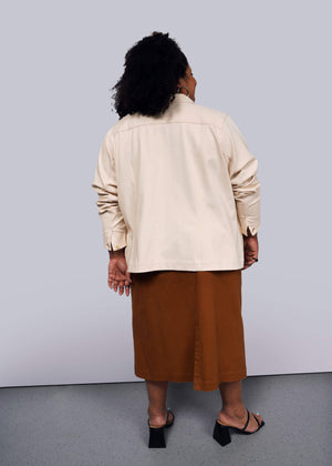 1X=A person with curly hair stands with their back to the camera in a beige long-sleeve shirt, brown skirt, and black open-toe heels. Completing the look is The Essential Denim Utility Jacket, featuring deep pockets. The background is plain white.