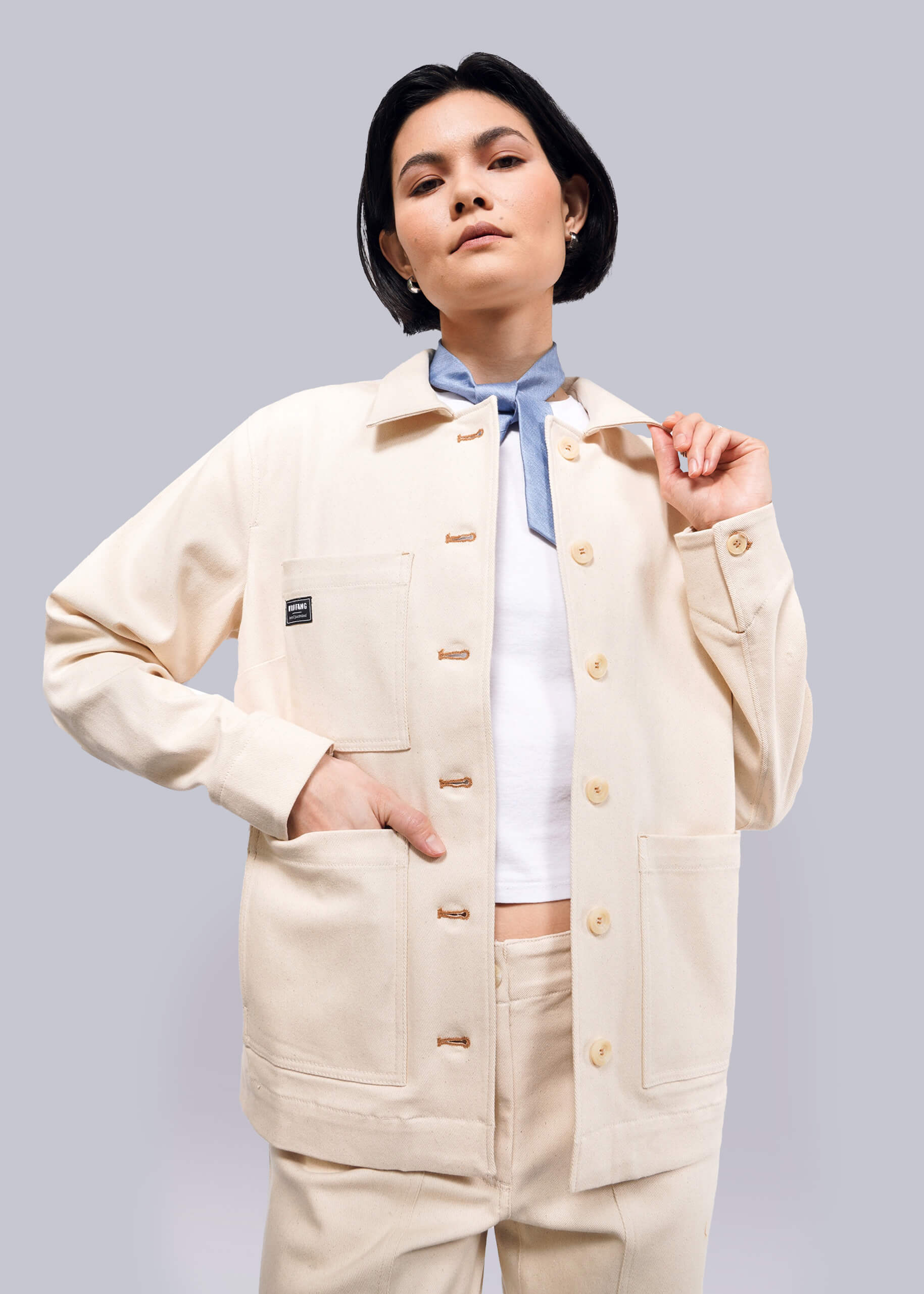 A person with short black hair wears The Essential Denim Utility Jacket in beige, featuring deep pockets over a white shirt. They pose confidently against a plain white background.
