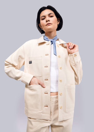 S=A person with short black hair wears The Essential Denim Utility Jacket in beige, featuring deep pockets over a white shirt. They pose confidently against a plain white background.