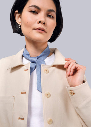 S=A woman with short black hair wears The Essential Denim Utility Jacket, featuring large buttons and deep pockets. She accessorizes with a blue neck scarf and small hoop earrings, holding the collar and looking slightly upward against a white background.