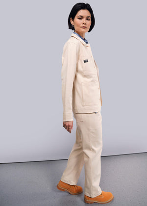 S=A person with short dark hair wearing The Essential Denim Utility Jacket paired with matching pants and tan shoes stands on a gray speckled floor against a light background. Deep pockets on the beige jacket combine style and stretch comfort as they look sideways.