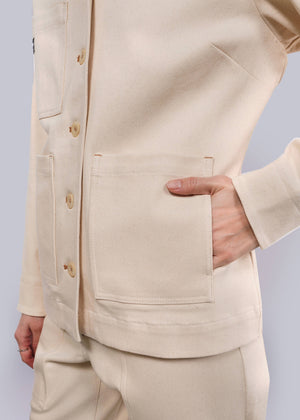 S=A person wears The Essential Denim Utility Jacket in beige with deep pockets and matching pants. Their right hand is in a pocket, showcasing textured fabric and light buttons for style and stretch comfort against a plain white background.
