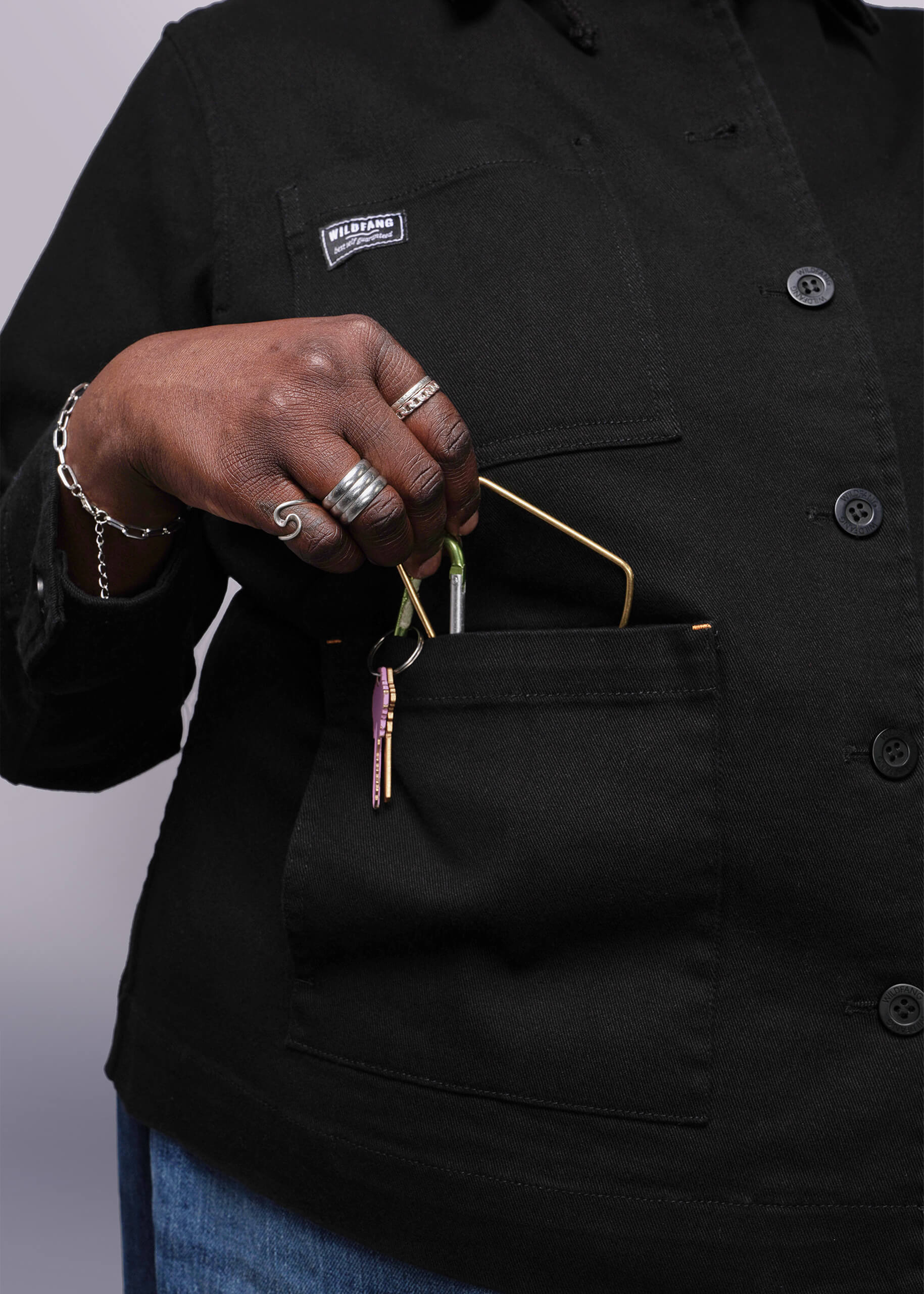 1X=Person wearing a black jacket with a Wildfang logo, placing keys in a pocket, on a light grey background