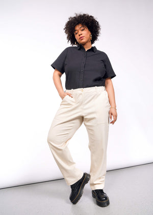 1X=A person with curly hair stands confidently against a plain backdrop, sporting a stylish black short-sleeve button-up shirt and The Essential Denim Utility Pant in cream. The pants feature an adjustable waist and pair perfectly with their black platform shoes, as they look forward, hand in pocket.