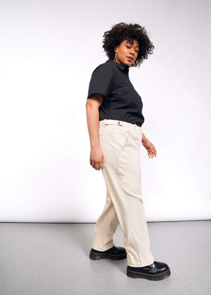 1X=A person with curly hair stands sideways, wearing a black t-shirt and The Essential Denim Utility Pant in beige with an adjustable waist. Black boots complete the look against a plain white backdrop and gray floor in an indoor setting.
