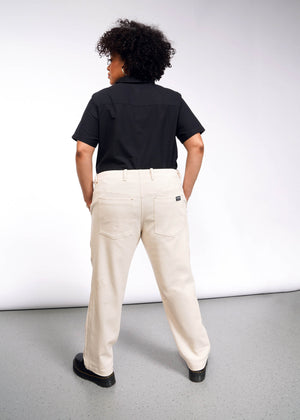 1X=A person with curly hair stands facing away from the camera, wearing a black shirt, The Essential Denim Utility Pant in light beige with an adjustable waist, and black shoes. The background is a simple white and gray studio setting.