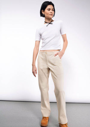 S=A person with short dark hair stands confidently in a white t-shirt, The Essential Denim Utility Pant in beige with an adjustable waist, and light brown boots. They wear a black choker necklace and have one hand in their pocket, against a plain white wall and gray floor.
