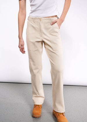 S=A person stands on a gray floor against a white background, wearing a white t-shirt and The Essential Denim Utility Pant in beige with an adjustable waist, wide legs, and large front pockets, paired with tan work boots.