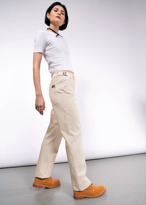 S=Someone in a white t-shirt, The Essential Denim Utility Pant, and tan shoes confidently poses against a plain white background. Their short, dark hair complements their casual yet stylish appearance, enhanced by the adjustable waist of the pants.